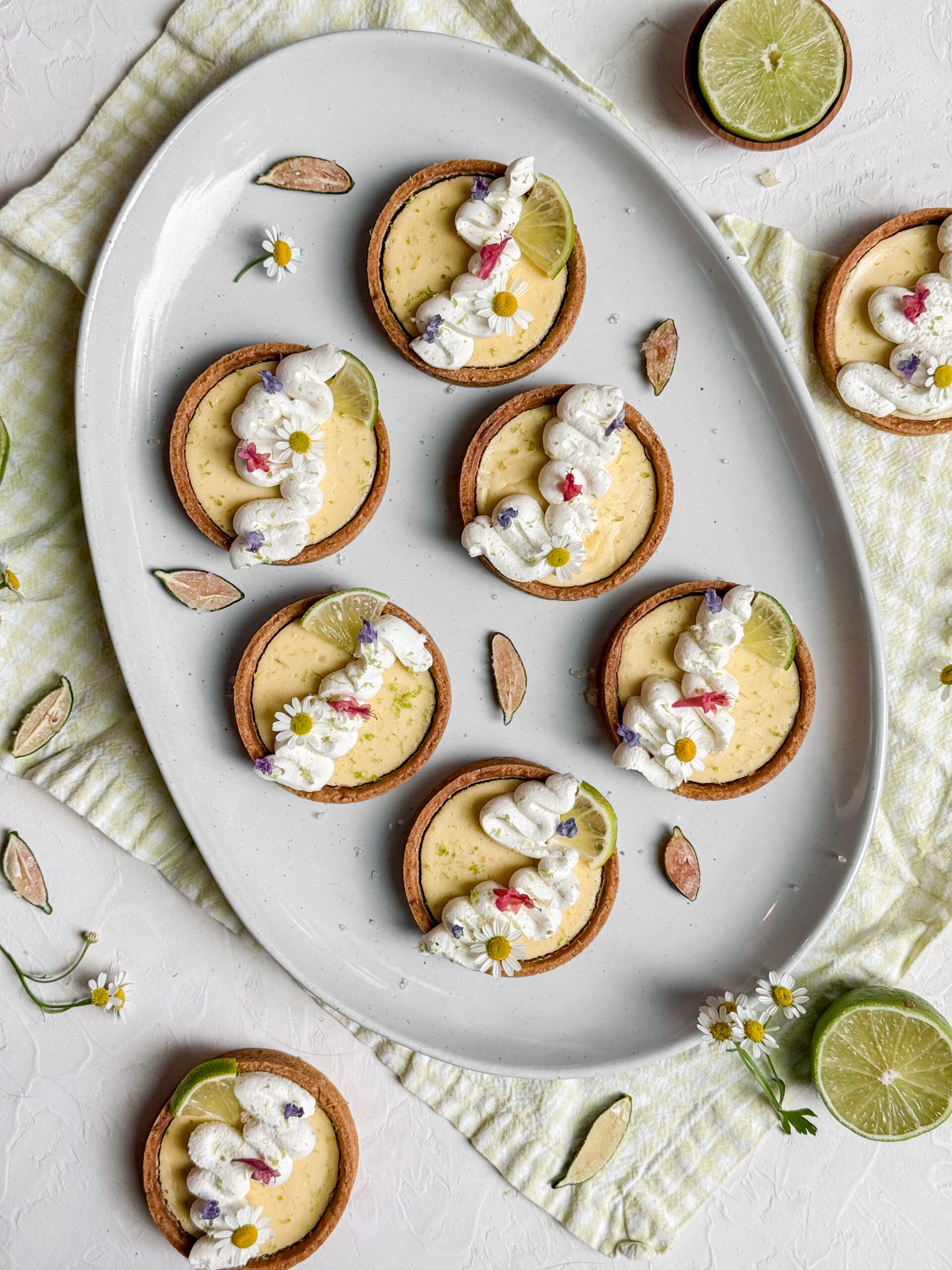 A plate of mini Key Lime Pies with whipped cream swirls, lime slices, and edible flowers, beautifully styled on a light background.